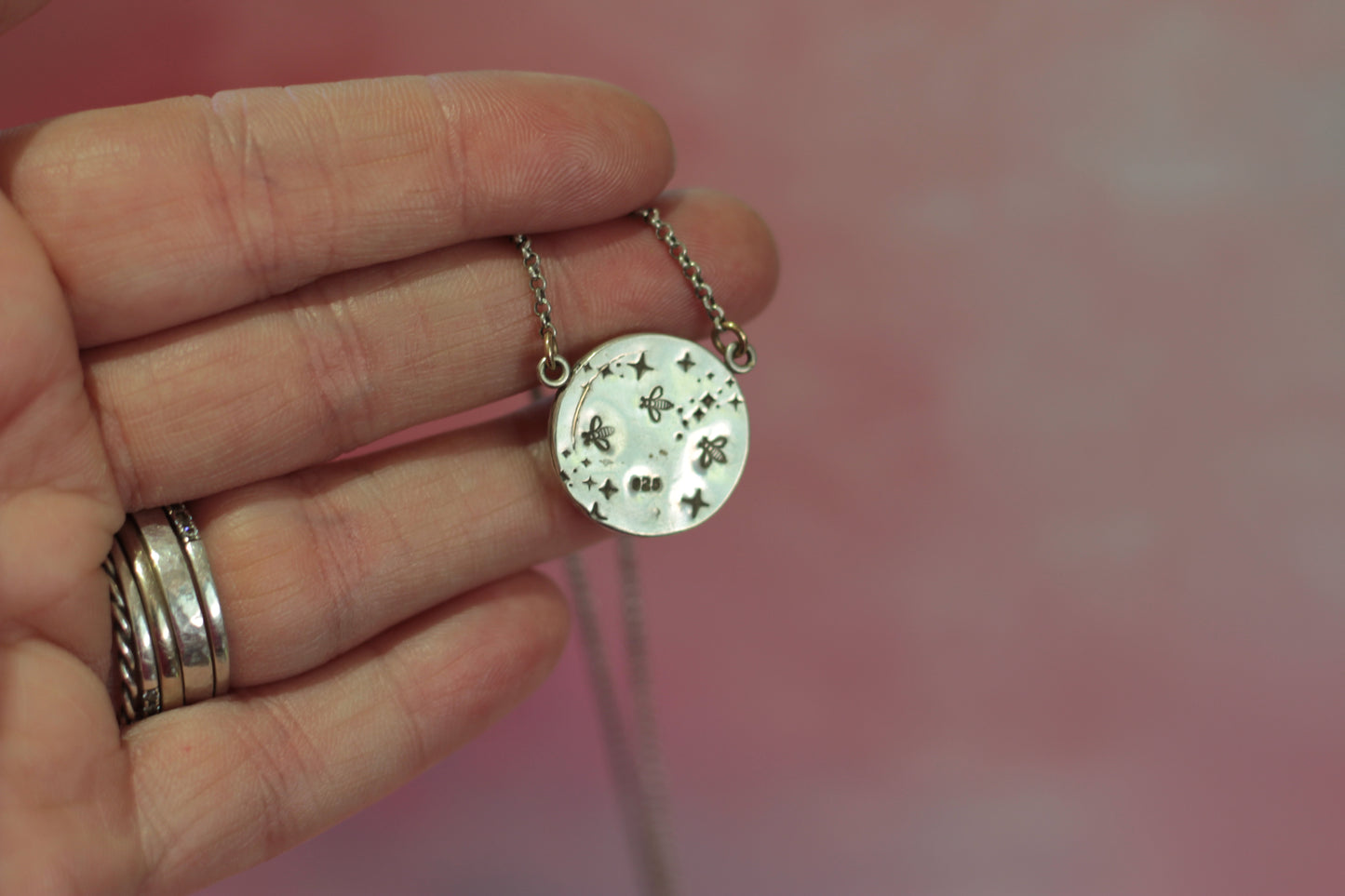 Hand holding a silver necklace with a round pendant against a pink background
