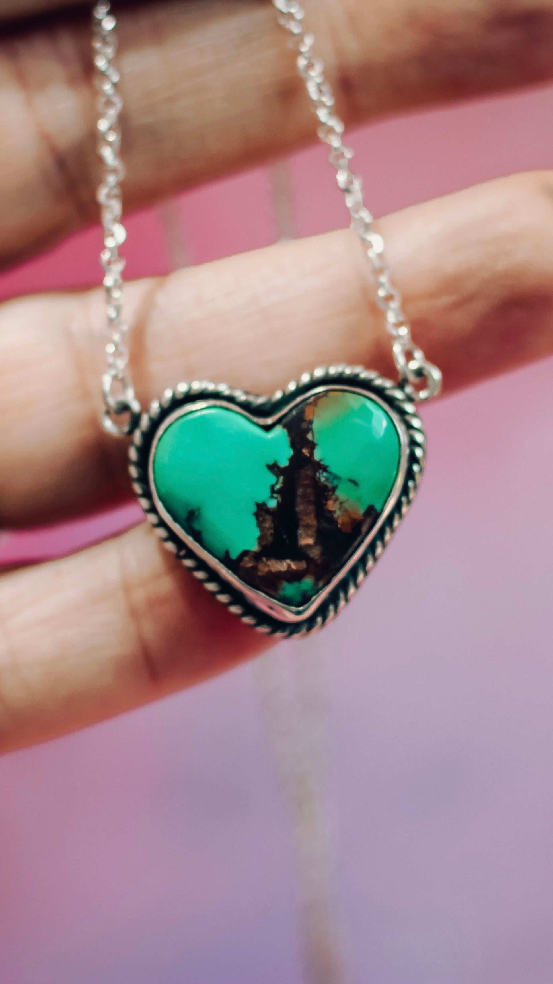 Heart-shaped turquoise pendant on a silver chain held by a hand against a pink background